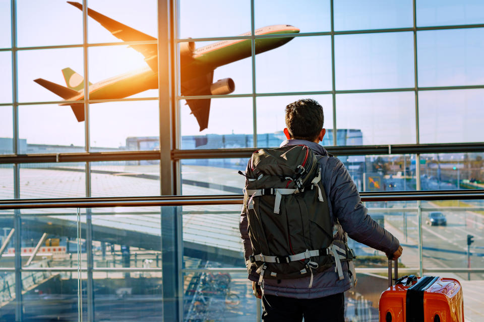 Travel concept Male tourist is standing in airport and looking at aircraft flight through window. He is holding tickets and suitcase. Sunset